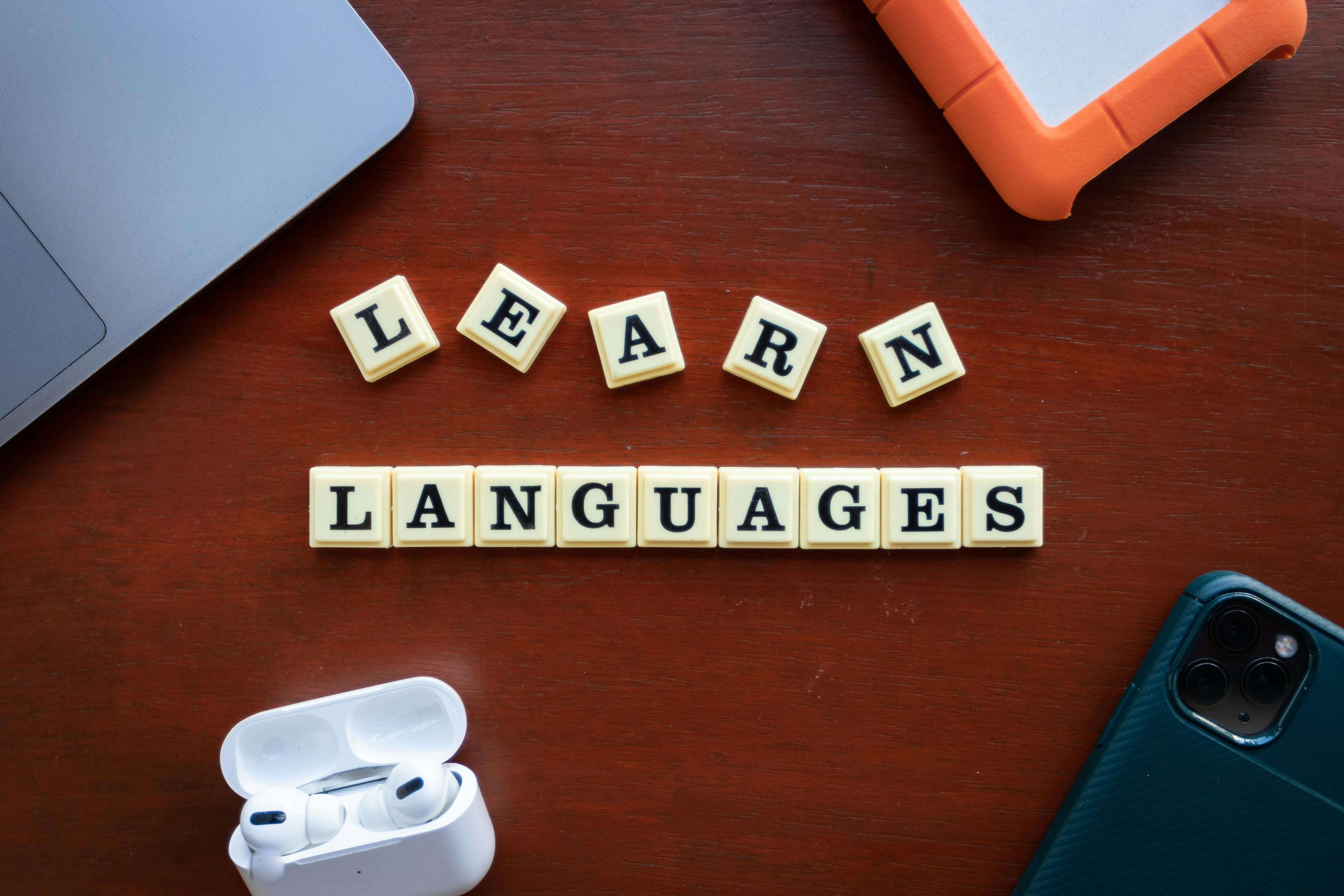 Motivace k učení cizích jazyků moderním způsobem Scrabble kostičky se slovem „Learn languages“, doplněné o bezdrátová sluchátka a mobil, symbolizují moderní a motivující přístup k učení cizích jazyků.