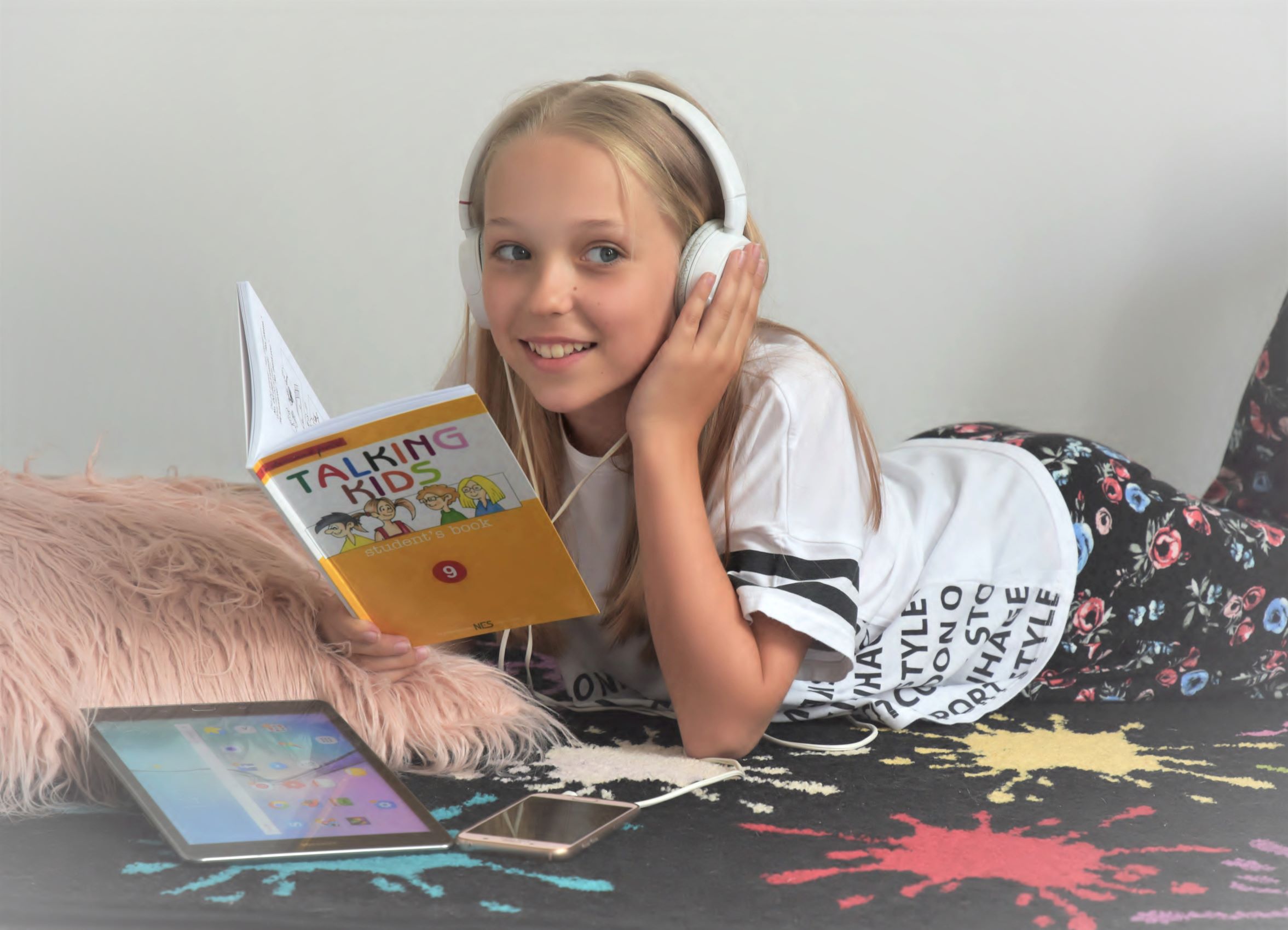 Zábavná angličtina pro děti s učebnicí Talking Kids Dívka se sluchátky leží na posteli a učí se angličtinu z učebnice Talking Kids, moderní a zábavná forma výuky angličtiny pro děti z pohodlí domova.